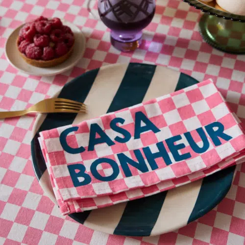 Set de 2 serviettes de table en coton, motif damier rose, message “Casa Bonheur” en situation sur une table dressée pour un repas familial