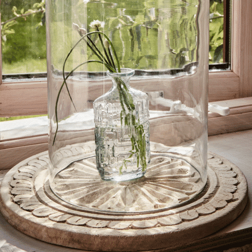 Plateau rond sur pieds en bois sculpté "Treasure" - Twicy Store disposé devant une fenêtre avec un bouquet de fleurs sous cloche