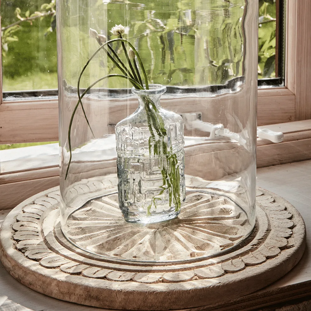 Plateau rond sur pieds en bois sculpté "Treasure" - Twicy Store disposé devant une fenêtre avec un bouquet de fleurs sous cloche
