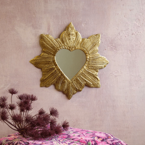 Miroir ex-voto en laiton doré "Coeur Royal" - Twicy Store suspendu à un mur de chaux rose devant une pile de tissu colorés et un bouquet mauve